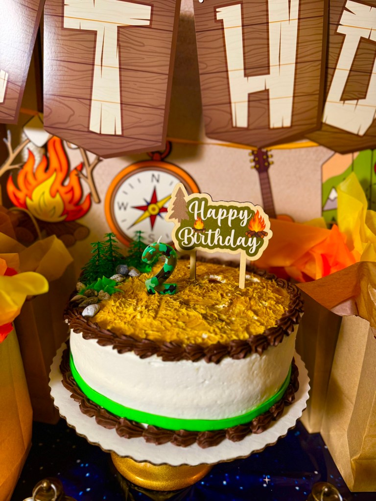 Close-up of camping birthday cake with textured frosting, mini trees, rocks, and number two candle.