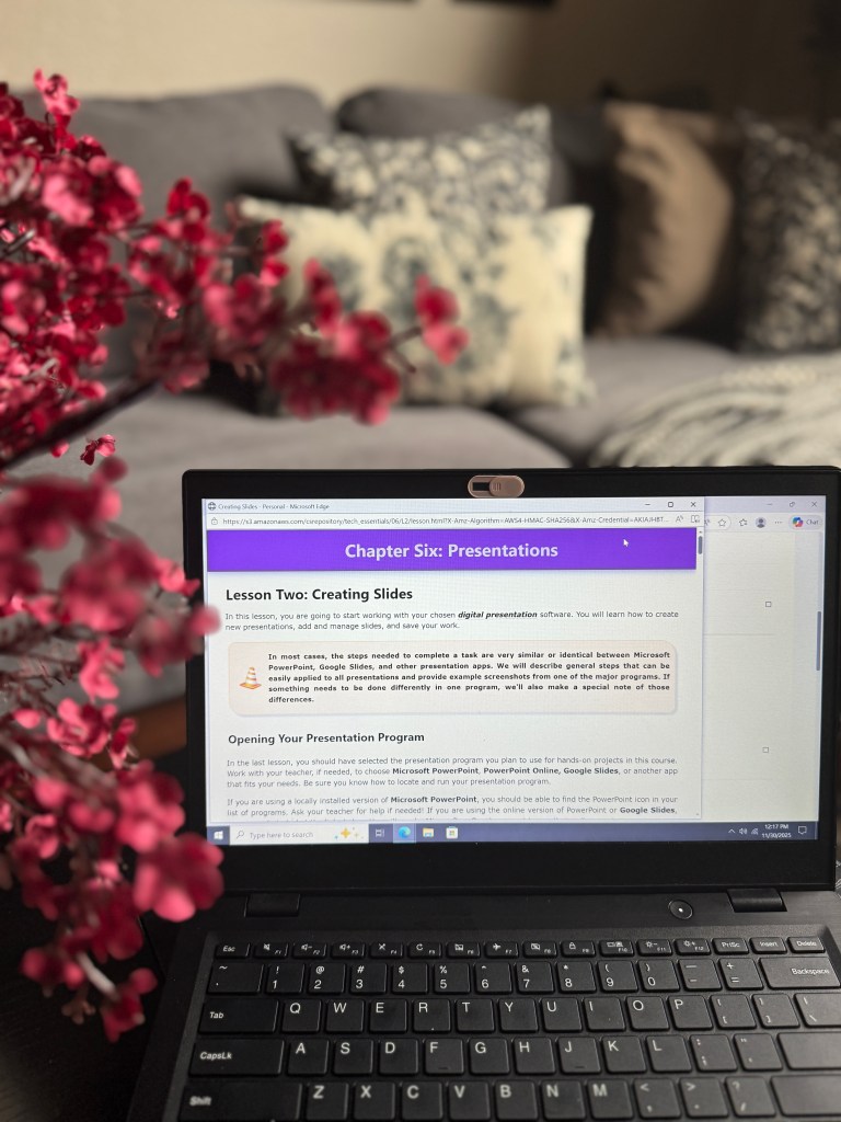 A laptop displaying a lesson on creating digital presentations, accompanied by a pink floral arrangement in the foreground. The lesson text discusses using presentation software like Microsoft PowerPoint and Google Slides.