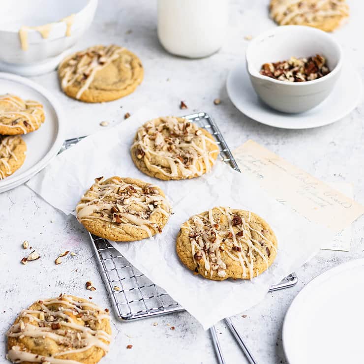 Close-up of freshly baked pumpkin cookies topped with a drizzle of icing and chopped pecans, arranged on a wire cooling rack and scattered with crumbs and ingredients.