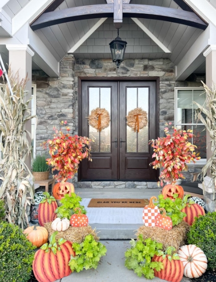 A welcoming fall porch with double wooden doors adorned with decorative wreaths, surrounded by colorful pumpkins, vibrant leaves, and festive decor creating an inviting autumn atmosphere.