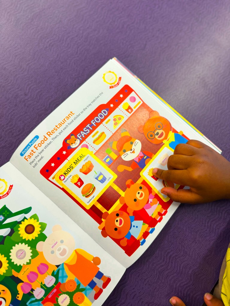 A child's hand placing a food sticker on a colorful page depicting a fast food restaurant scene with cartoon animals and food items.