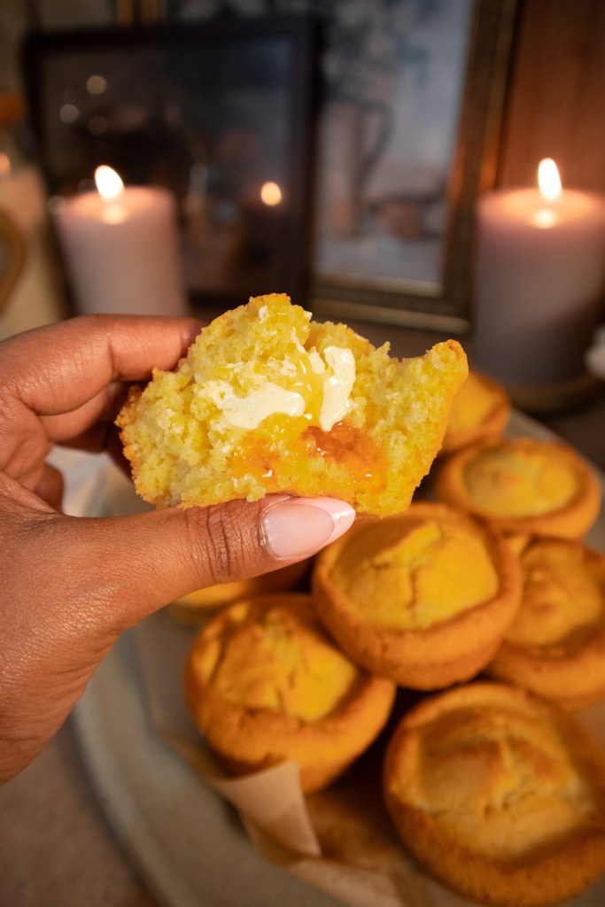 A hand holding a vegan cornbread muffin with melted butter inside, with more muffins in the background and lit candles.
