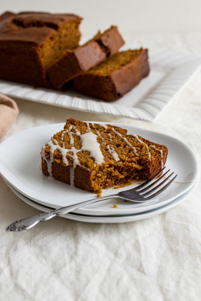 A slice of pumpkin bread drizzled with a white glaze, served on a plate with a fork, alongside a loaf of pumpkin bread in the background.