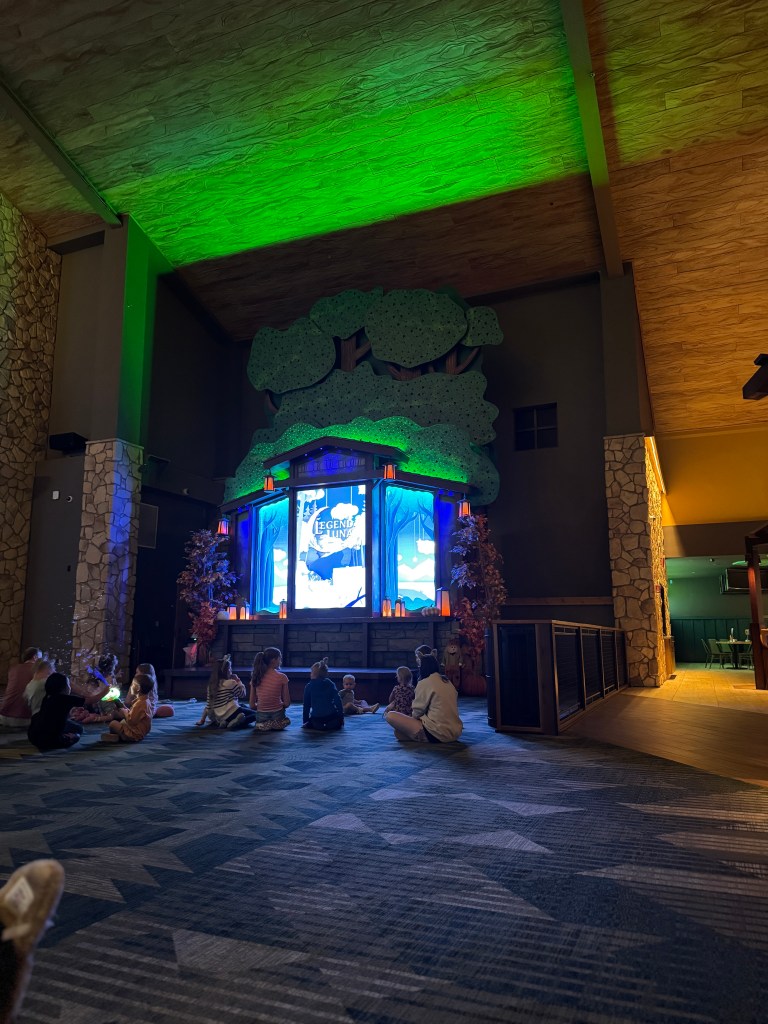 Children gathered on the floor of the Great Wolf Lodge lobby for a storytelling session, surrounded by colorful decorations and a screen displaying animated visuals.