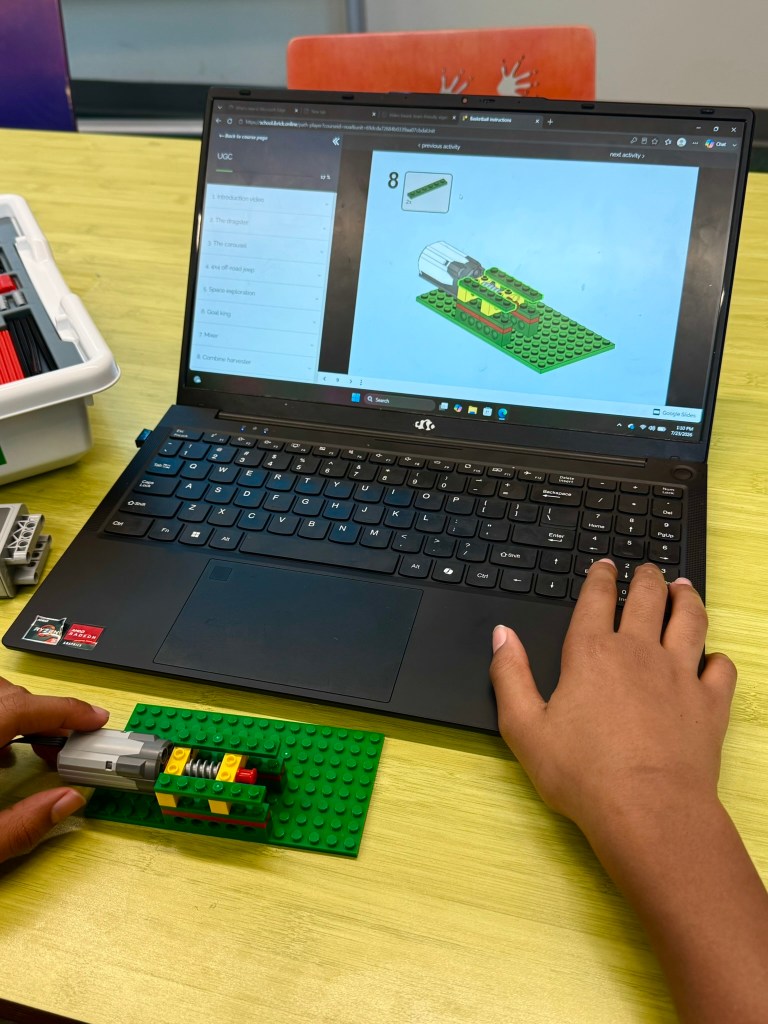 A person assembling a LEGO project next to a laptop displaying step-by-step instructions for building on a green LEGO baseplate.
