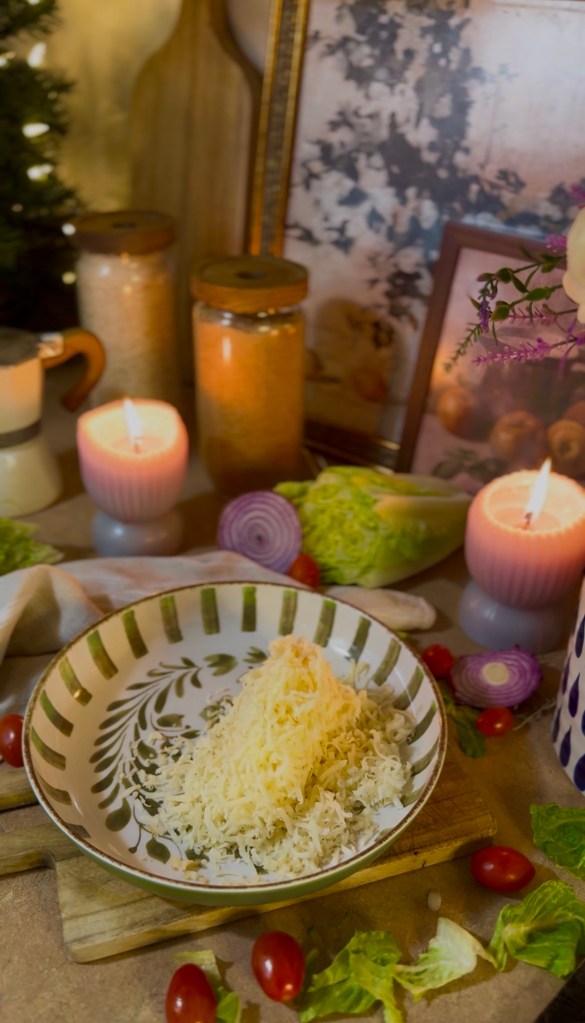A bowl with grated cheese on a wooden board, surrounded by fresh vegetables, candles, and decorative items.