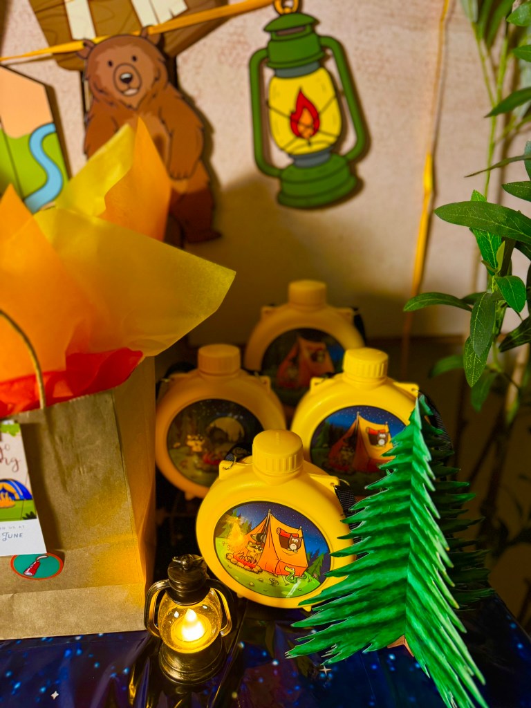 Close-up of camping birthday party table decor with a yellow canteen, toy binoculars, fire napkins, and wood-look cups.