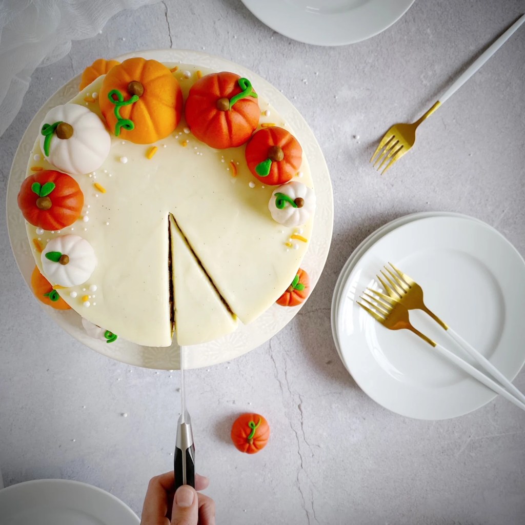 A beautifully decorated cake with a smooth white icing and fondant pumpkins on top, being sliced with a cake server, surrounded by white plates and gold forks.