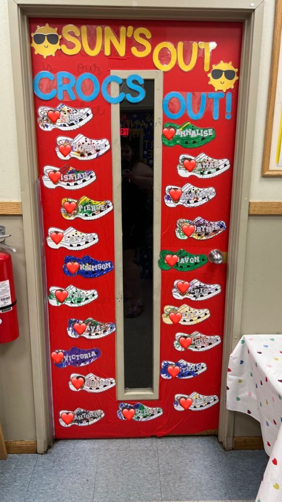 Brightly colored door decorated with a 'SUN'S OUT, CROCS OUT!' sign, featuring multiple images of Crocs shoes and heart symbols, with names displayed beside each shoe.