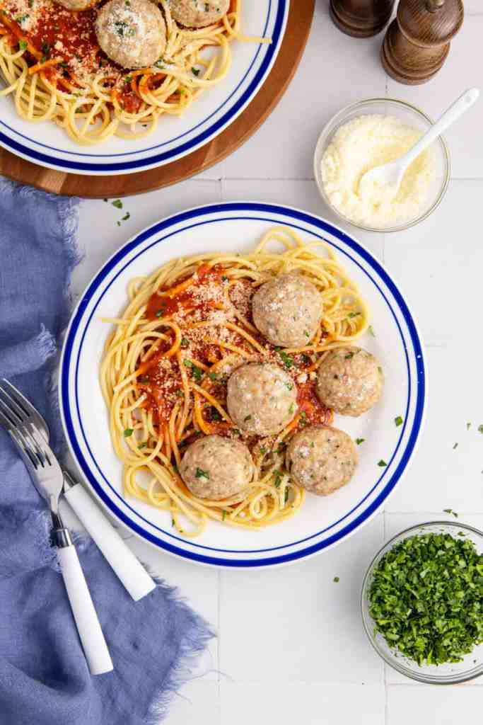 A plate of spaghetti topped with ground chicken meatballs in marinara sauce, grated cheese, and chopped herbs, accompanied by a small bowl of parmesan cheese and a sprinkle of fresh parsley.