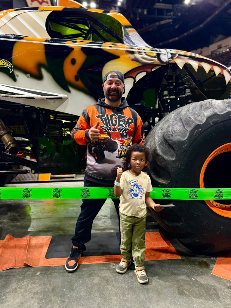 Young child poses with the Tiger Shark driver beside the giant monster truck tire.