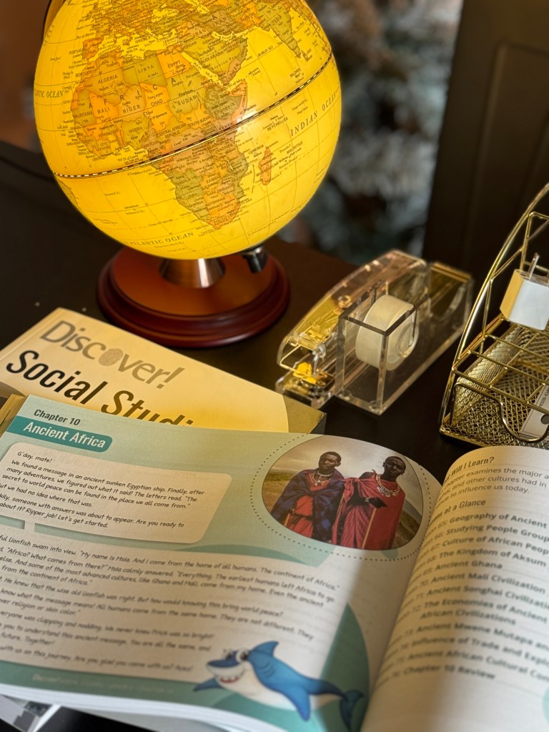 A glowing globe focusing on Africa sits on a dining table beside an open book titled 'Discover! Social Studies' with illustrations and text about Ancient Africa.