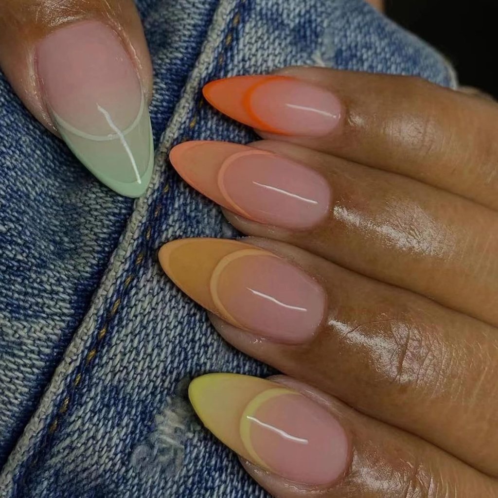 A close-up of a hand with stylish, gradient nail designs featuring pastel colors, including mint green, orange, and yellow tips, against a denim background.