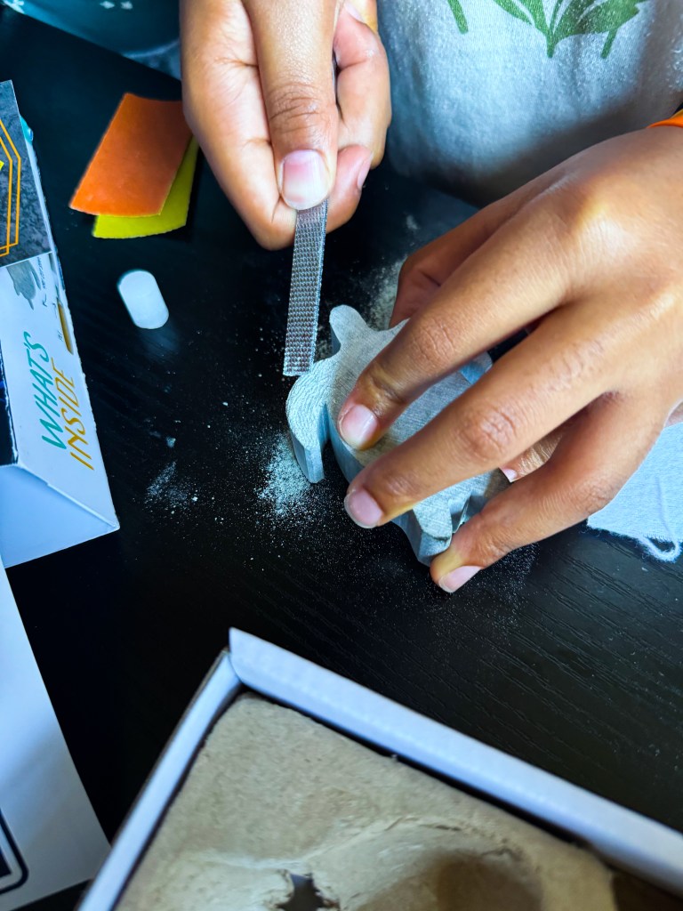 A person's hands using a file to shape a small object, with colorful crafting materials and dust visible on a black surface.