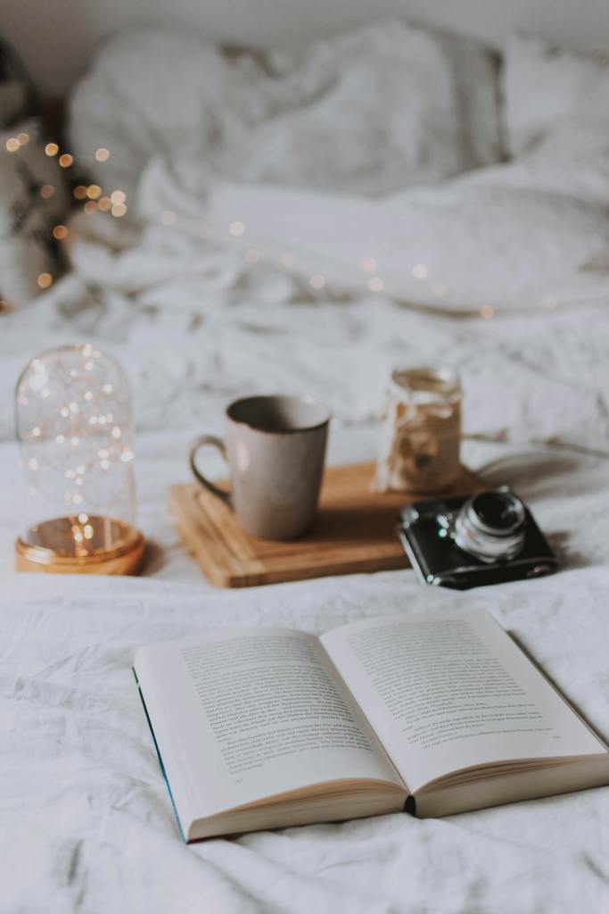 A cozy bedroom scene featuring an open book on a white bedspread, a warm cup of coffee, a glass jar, and a camera on a wooden tray, with soft fairy lights in the background.